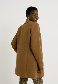 Abrigo de mezcla de lana marrón con un diseño estructurado, que incluye un cuello y bolsillos, combinado con pantalones de estampado de leopardo. Textura suave y ajuste entallado.