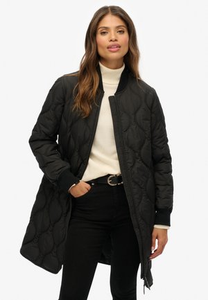 Vrouw in een lange zwarte gewatteerde jas over een crèmekleurige turtleneck en zwarte broek, poserend met één hand in haar zak tegen een witte achtergrond.