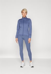 Blå atletisk utstyr bestående av en glidelåsjakke og tettsittende leggings, laget av glatt stoff. Joggesko fullfører antrekket.
