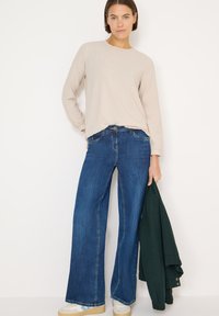 Jasnobeżowa bluzka z długim rękawem i marszczonym dekoltem, skomponowana z ciemnoniebieskimi, szerokimi spodniami jeansowymi. Modelka trzyma zieloną kurtkę, widoczne brązowe buty.