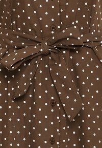 Material maro cu buline albe, curea de talie legată într-un arc și nasturi vizibili de-a lungul centrului.