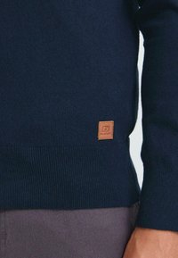 Tamnoplavi pleteni pulover s malim smešnim kožnim logotipom blizu rubа, nošen preko tamnosivih hlača.