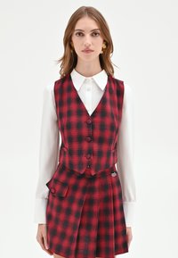 Gilet a quadri rossi e neri con cinque bottoni, abbinato a una gonna plissettata. Camicia bianca con colletto sotto, con dettaglio della tasca laterale.