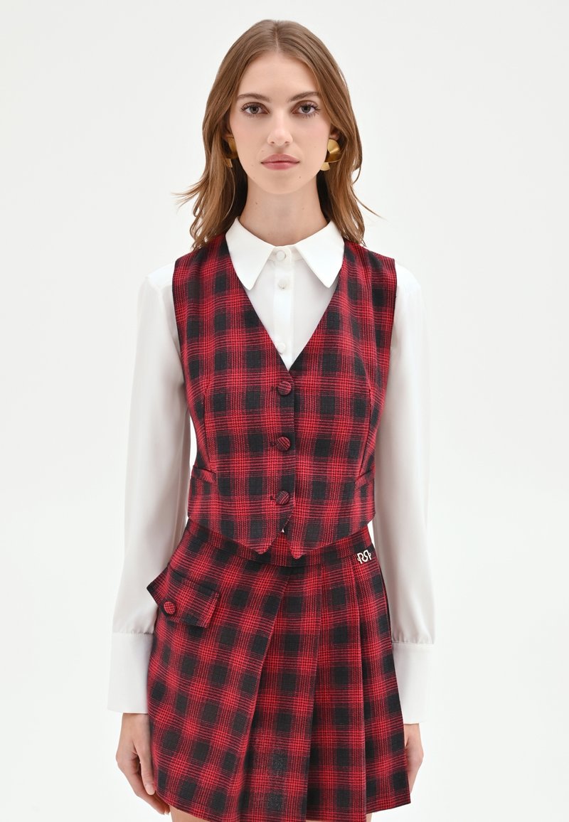 Gilet a quadri rossi e neri con cinque bottoni, abbinato a una gonna plissettata. Camicia bianca con colletto sotto, con dettaglio della tasca laterale.