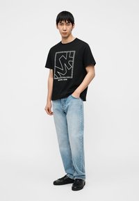 Μαύρο βαμβακερό T-shirt με λευκό τυπωμένο λογότυπο, σε συνδυασμό με τζιν σε απευθείας γραμμή ανοικτού μπλε χρώματος και μαύρα παπούτσια. Απλός, casual σχεδιασμός.