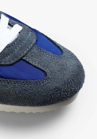 Zapatilla con parte superior de ante gris oscuro y detalles en tela azul profundo. Presenta cordones blancos y una suela texturizada para mayor agarre.