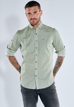 Man with tattoos wearing a patterned light green long-sleeve shirt with rolled-up sleeves and dark jeans, standing against a plain light background.