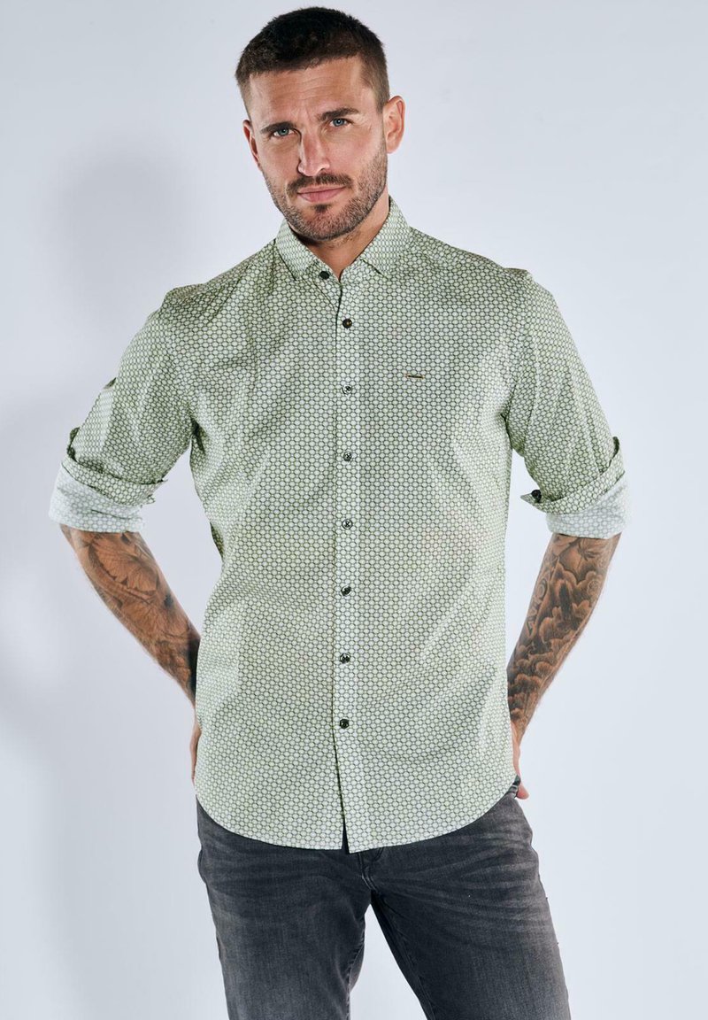 Mann mit Tätowierungen, der ein gemustertes, hellgrünes Langarmshirt mit hochgekrempelten Ärmeln und dunklen Jeans trägt, steht vor einem schlichten, hellen Hintergrund.