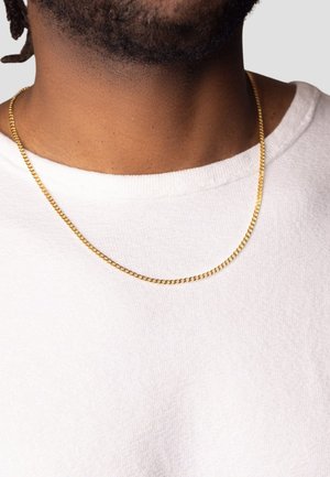 Goldene Halskette mit gedrehten Gliedern, die auf einem nackten Hals ruht, kombiniert mit einem strukturierten weißen Hemd. Glatte Oberfläche und glänzendes Finish.
