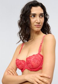 Γυναίκα με σγουρά σκούρα μαλλιά που κοιτάζει δεξιά, φορώντας ένα λεπτομερώς διακοσμημένο κόκκινο δαντελένιο σουτιέν με κεντήματα λουλουδιών, με τα χέρια σταυρωμένα πάνω σε ουδέτερο φόντο.