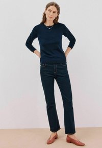 Granatowy wełniany sweter z długimi rękawami, zestawiony z ciemnymi dżinsami. Model nosi jasnoróżowe płaskie buty; widoczny minimalistyczny naszyjnik.