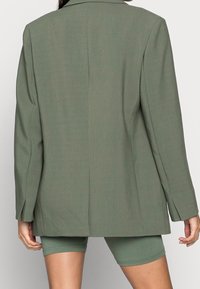 Blazer vert olive à coupe ample, avec une fente unique dans le dos et des revers nets. Porté sur un short de cycliste vert assorti, mettant en valeur un look superposé.
