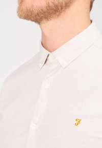 Camisa de lino color gris claro con botones, que presenta un pequeño logo amarillo bordado en el pecho y botones blancos. Superficie texturizada.