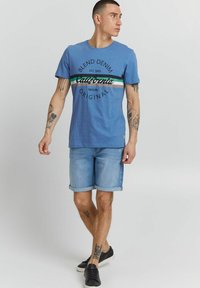 Zils kokvilnas T-krekls ar melnu uzrakstu "Blend Denim California". Valkāts ar gaiši ziliem džinsu šortiem un melnām sportakurpēm, parādot tetovējumus uz rokām.