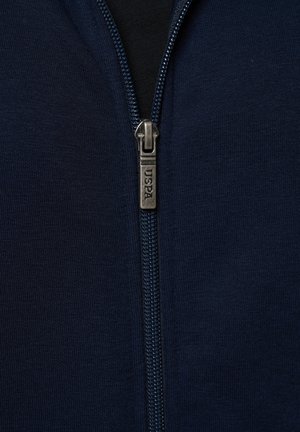 Detaljni prikaz tamnoplave tkanine munje sa srebrnim povlačnim dijelom na kojem je ugraviran natpis "USPA" na tamnom odjevnom predmetu.