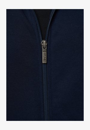 Detaljni prikaz tamnoplave tkanine munje sa srebrnim povlačnim dijelom na kojem je ugraviran natpis "USPA" na tamnom odjevnom predmetu.