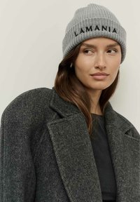 Egy nő szürke kötött sapkát visel, rajta a "LAMANIA" felirattal, sötétszürke halszálkás kabátban és arany fülbevalóban, kissé oldalra néz.