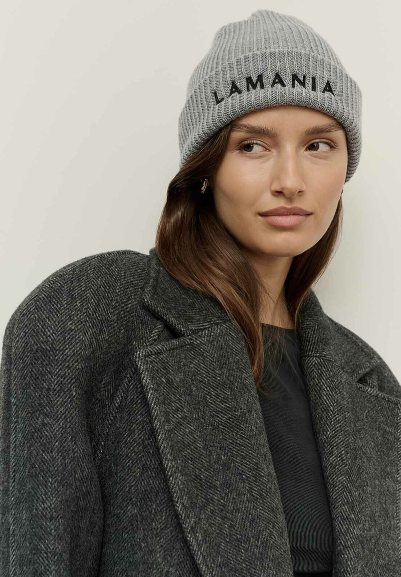 Egy nő szürke kötött sapkát visel, rajta a "LAMANIA" felirattal, sötétszürke halszálkás kabátban és arany fülbevalóban, kissé oldalra néz.