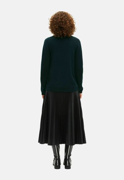 Mørkegrøn sweater med rund halsudskæring, parret med en plisseret sort nederdel, der går til midten af læggen, og glansfulde sorte støvler med spænder.