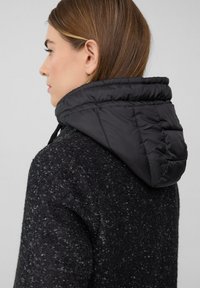 Vrouw met bruin haar, gekleed in een gestructureerde zwarte jas en een pluchen zwarte capuchon, kijkt naar links tegen een eenvoudige lichtgrijze achtergrond.