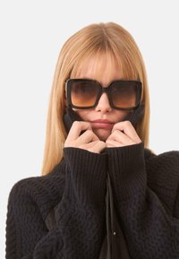 Suéter negro acanalado con cuello alto, combinado con gafas de sol grandes y cuadradas con lentes marrones degradados. Cabello rubio claro.
