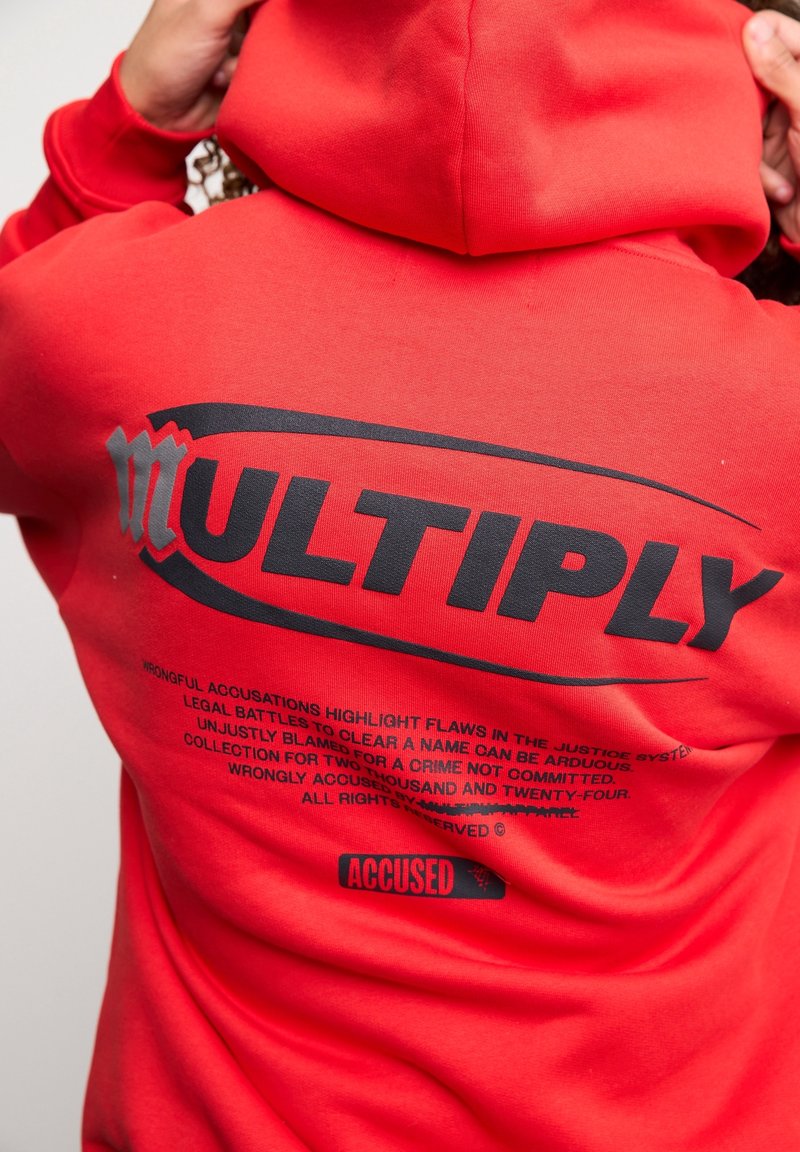 Persona con un hoodie rosso con grande testo nero "MULTIPLY" e un testo più piccolo riguardo le accuse ingiuste e le battaglie legali sul retro.