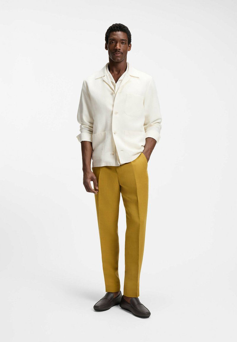 Homme portant une veste blanche boutonnée, un pantalon jaune moutarde et des chaussures marron foncé, debout avec une main dans la poche.