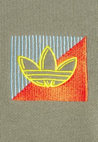 Vyšívání žlutého loga na zelené tkanině, s živým červeným a modrým geometrickým pozadím s jemnými vertikálními pruhy.