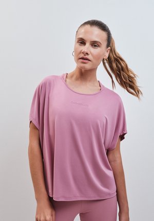 Frau mit heller Haut und braunen Haaren, zu einem Pferdeschwanz gebunden, trägt ein lockeres rosa Kurzarm-Top und passende Leggings vor einfarbigem Hintergrund.