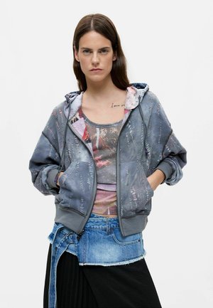 Jeune femme aux longs cheveux bruns portant un sweat à capuche zippé bleu usé, un débardeur imprimé, une jupe en jean par-dessus et une jupe noire, les mains dans les poches.