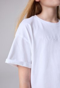 Witte katoenen T-shirt met opgerolde korte mouwen, met subtiele reliëfletters op de voorkant, een gladde textuur en een standaard ronde hals.
