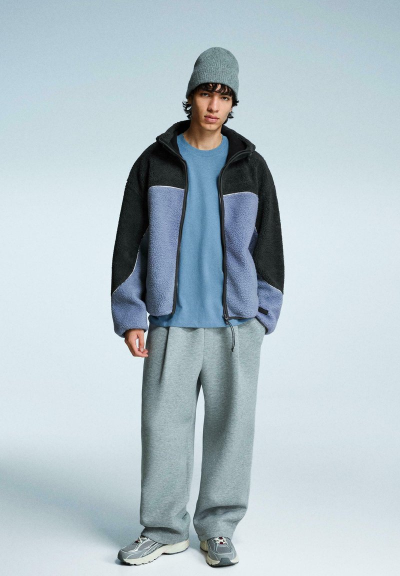 Fleecejack in zwart en paars, gecombineerd met een lichtblauwe T-shirt, grijze joggingbroek en grijze sneakers. Accessoires omvatten een grijze beanie.