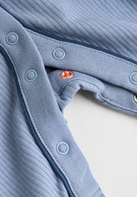 Helleblaue gerippte Stoff mit Druckknöpfen, versehen mit einem dunkelblauen Besatz und einem kleinen orangefarbenen Akzentknopf in der Nähe des Halsausschnitts.