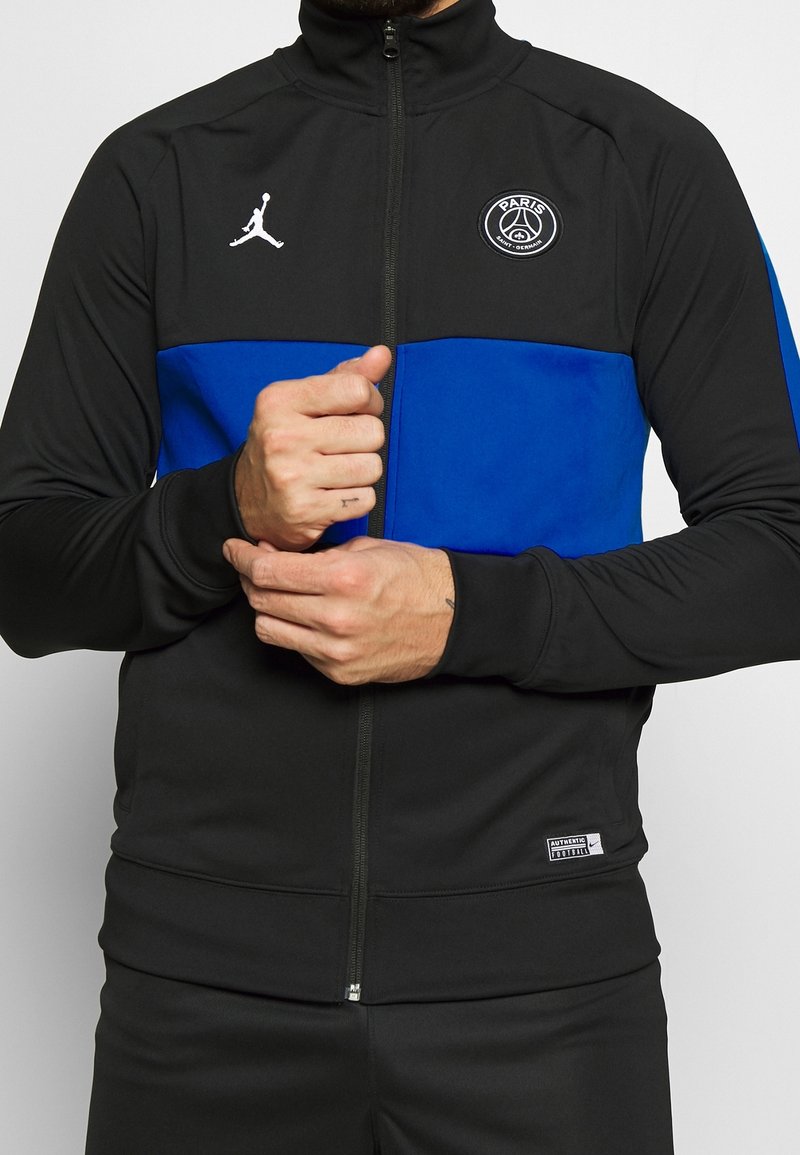 Homme portant une veste de sport noire et bleue Paris Saint-Germain avec fermeture éclair et logo Jumpman blanc, ajustant les poignets.