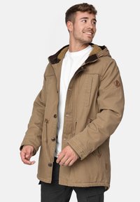 Veste en coton marron avec une capuche, dotée d'un col en peluche, de plusieurs poches avant et de fermetures à boutons. Tissu texturé et durable.