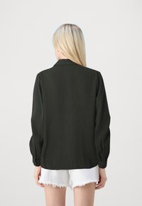 Blusa lunga verde scuro con collo a camicia e polsini elasticizzati, in tessuto testurizzato, abbinata a pantaloni corti bianchi sfilacciati.
