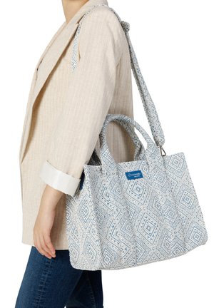 Femme portant un blazer beige et un jean bleu, tenant un grand sac fourre-tout blanc et bleu à motifs, avec des poignées courtes et une longue bandoulière.