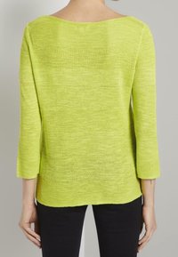 Haut en tricot vert citron avec manches trois-quarts, encolure dégagée et rayures texturées. Tissu léger et semi-transparent. Porté avec un pantalon noir.