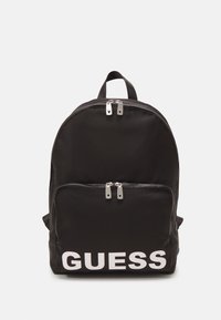 Zwarte nylon rugzak met het witte "GUESS" logo. Heeft een voorkant met ritsvak en dubbele ritslussen. Rond ontwerp met gewatteerde schouderbanden.