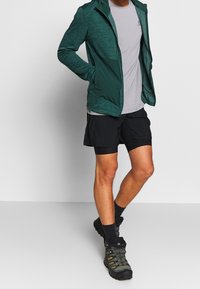 Grön texturerad jacka, grå crew-neck tröja, svarta shorts och grå vandringsskor. Noterbara färger och designinslag för aktivkläder.