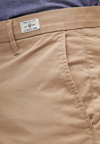 Khaki chino pants with a smooth texture, featuring a label near the waistband, and two side pockets. Subtle stitching details.