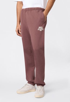 Osoba nosząca brązowe spodnie dresowe z elastycznym pasem i napisem „North Atlantic” na udzie oraz białe sneakersy, stojąca na tle jednolitego tła.