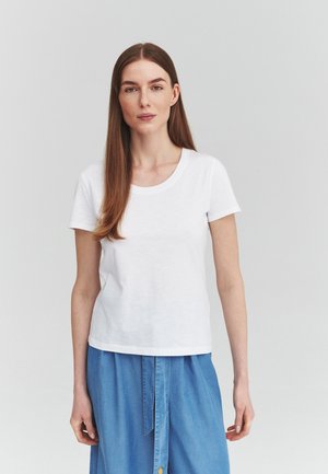Ung kvinde med langt brunt hår iført en almindelig hvid t-shirt og blå denim nederdel, der står mod en grå baggrund.