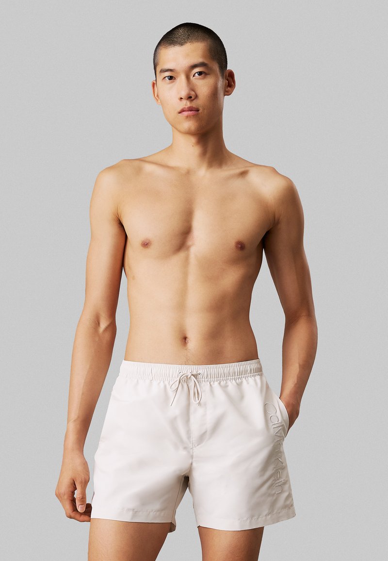 Shorts de bain beiges avec une taille élastique et un cordon de serrage ; dotés d'un logo subtil brodé sur le côté. Tissu lisse et léger.