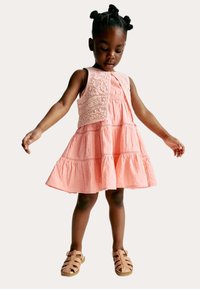 Robe rose avec une jupe à volants et des détails en dentelle élaborés sur le corsage, associée à des sandales marron clair. Texture lisse dans l'ensemble.