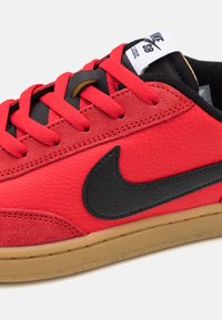 Sneaker in pelle rossa e suede con logo Nike nero, lacci neri e suola in gomma color cuoio. Presenta materiali testurizzati e dettagli cuciti.