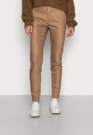 Personne portant un pantalon beige slim, des baskets blanches et un pull en maille marron, debout devant un fond blanc uni.