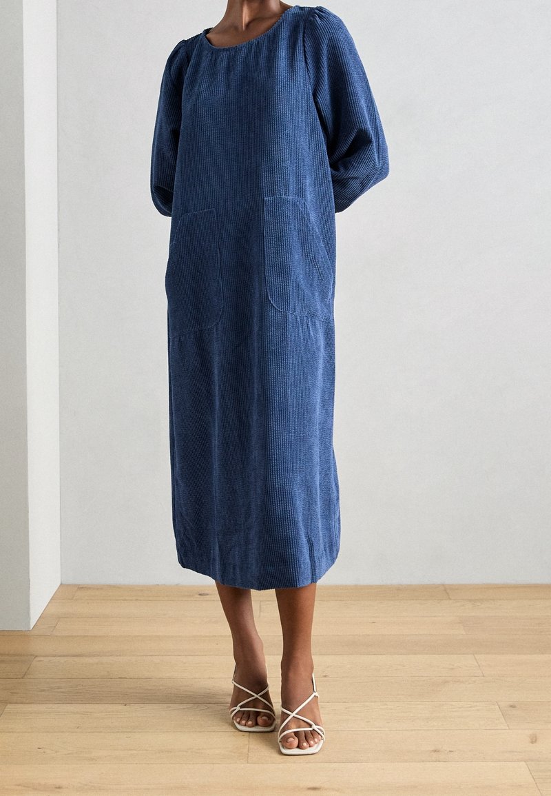 Femme portant une robe midi bleue ample à manches longues avec deux poches avant et des sandales blanches à talons hauts à brides, debout sur un sol en bois.