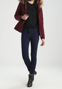 Bordeauxrød quiltet jakke med pelsforet hætte, sort top og tætsiddende mørkeblå jeans, kombineret med sorte ankelstøvler.