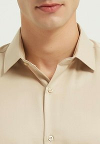 Chemise beige à col classique, avec une poche poitrine unique et un tissu lisse et texturé. Présente des coutures légères et des boutons en plastique.